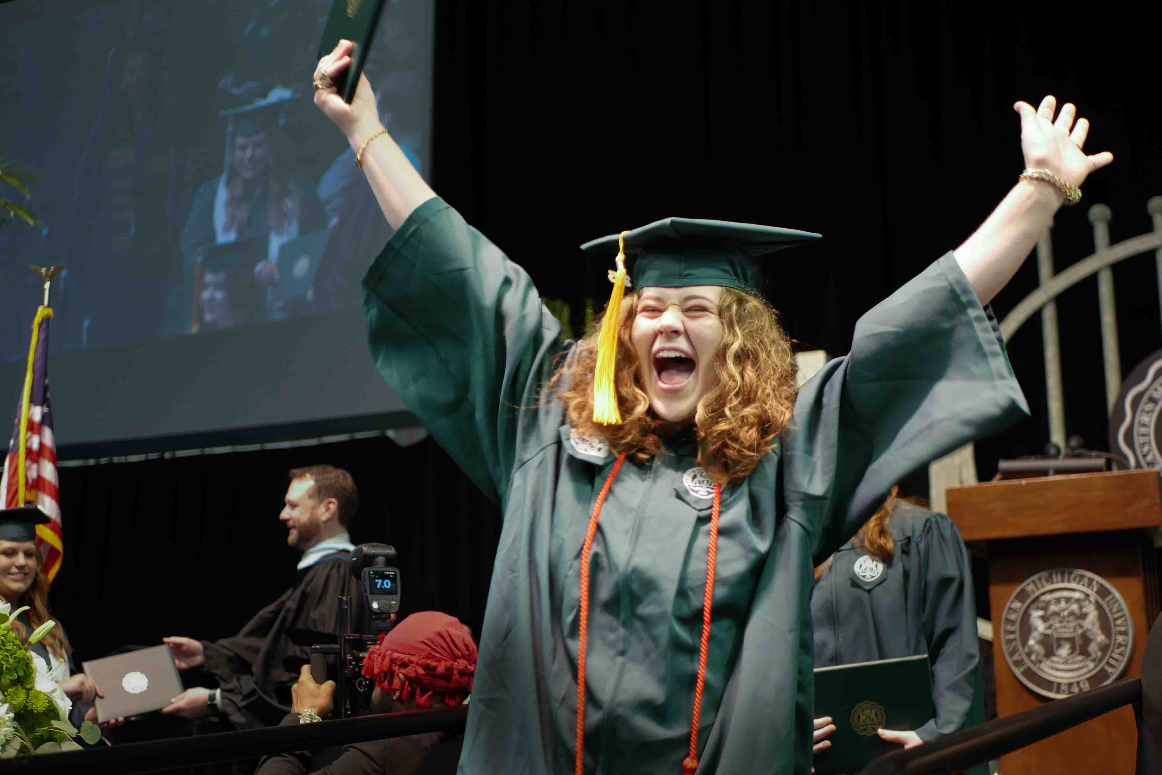 Kasey Deschler wearing a green gown throwing her arms up on the graduation stage.