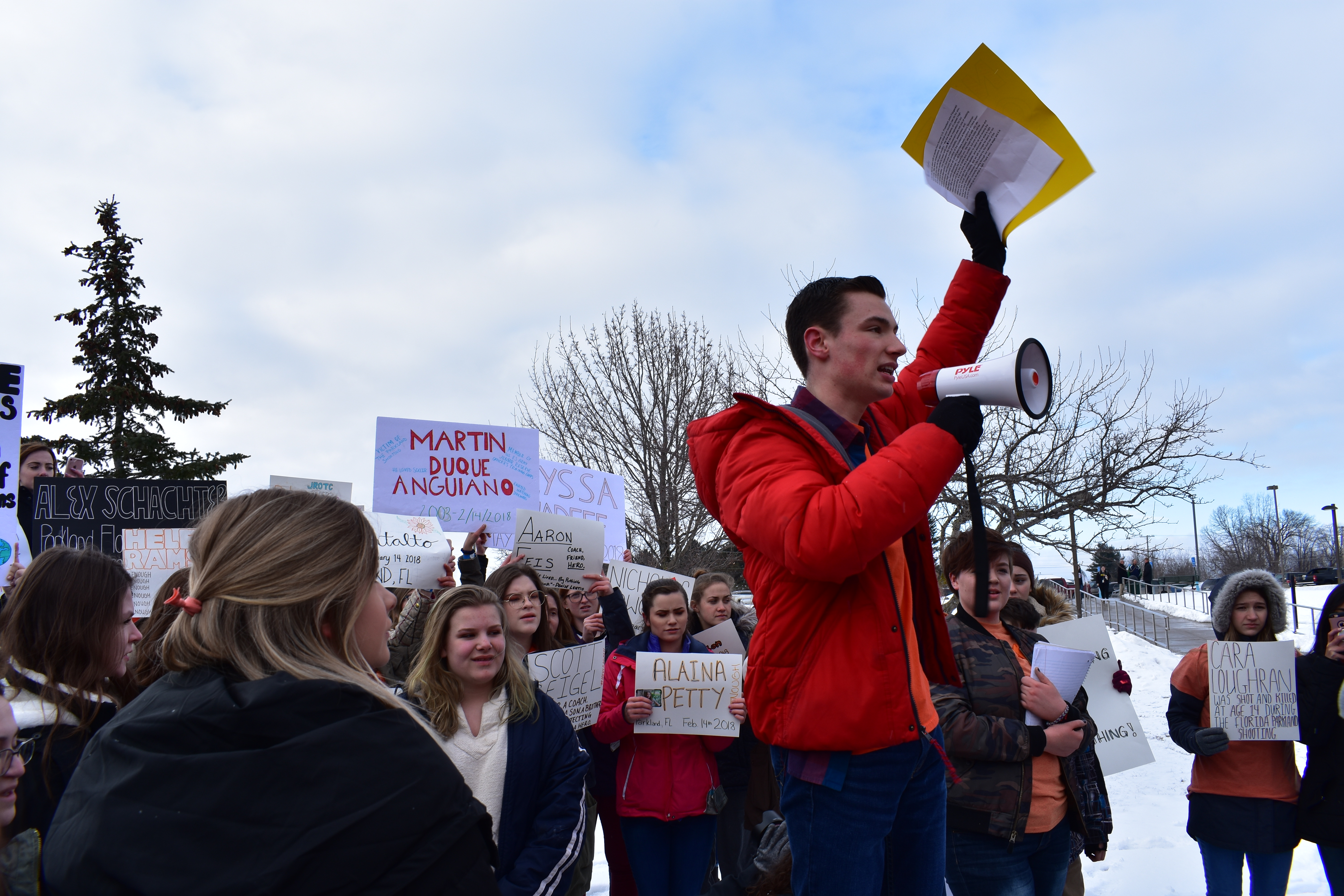 High school student Porter Hughes speaking into a megaphone with papers held high above his head. He is adressing a crowd of Lapeer High School students holding signs.